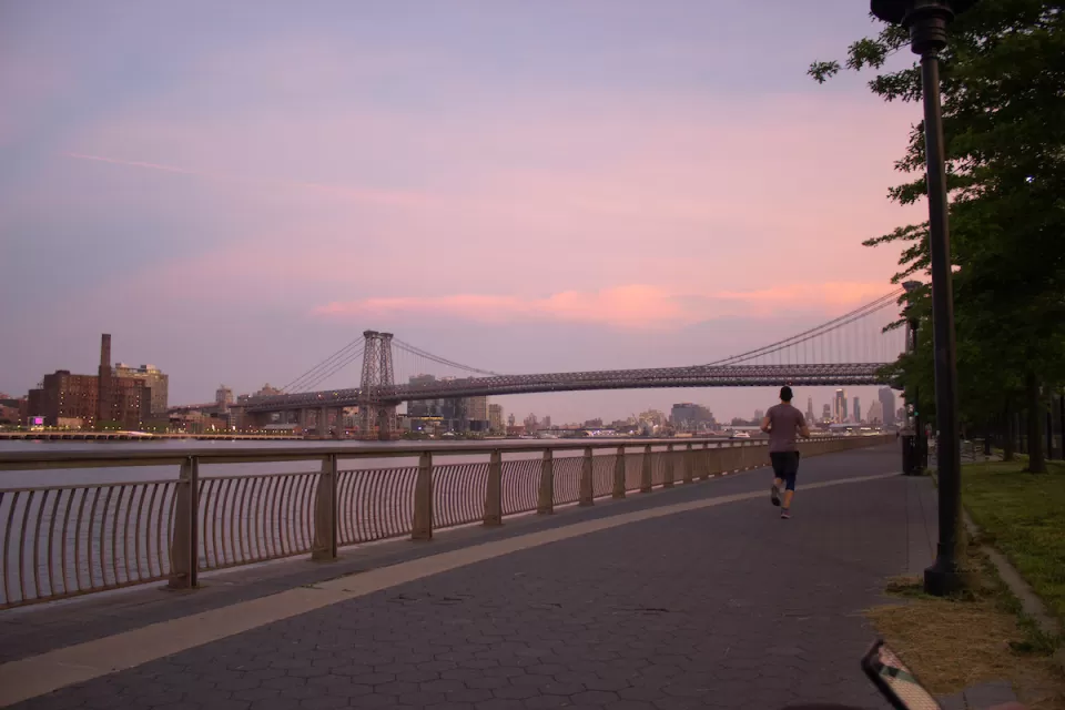 Sunset view of Williamsburg Bridge from East River Park walkway next to river with one jogger.